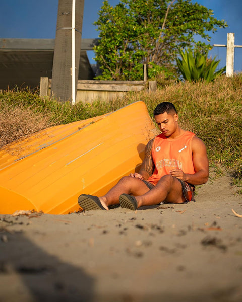 Man wearing Aotearoa basketball singlet with Paua design, sitting on a beach next to a yellow kayak